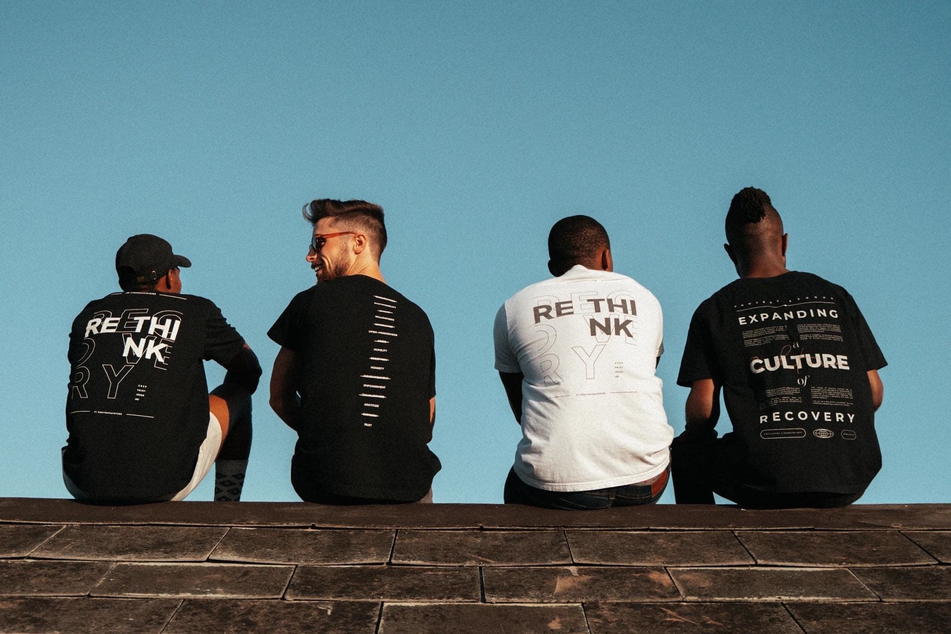 Four people sitting on a sea wall with their backs to the camera, each wearing a different design from the line