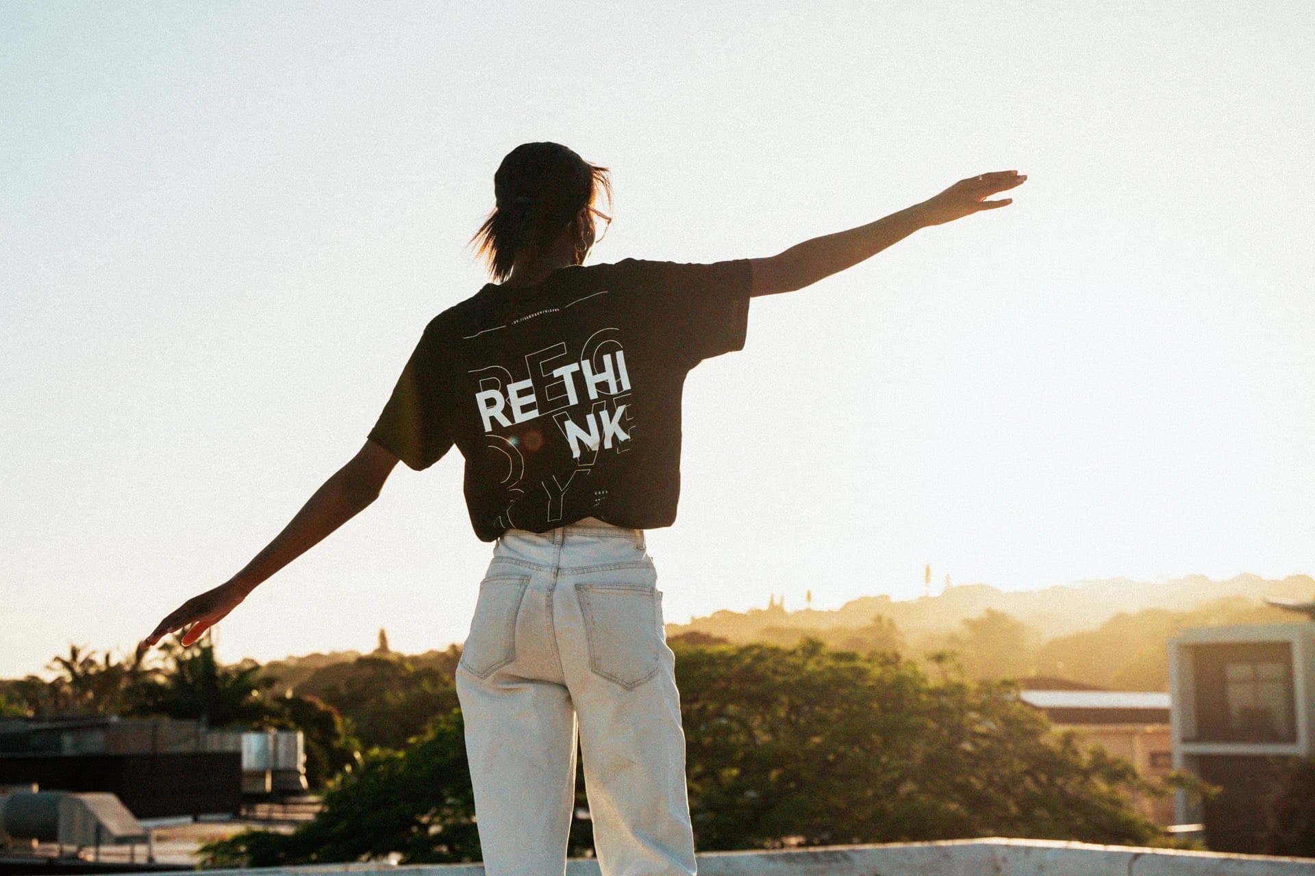 A woman on a rooftop at sunset, arms wide, RETHINK across the back of her tee