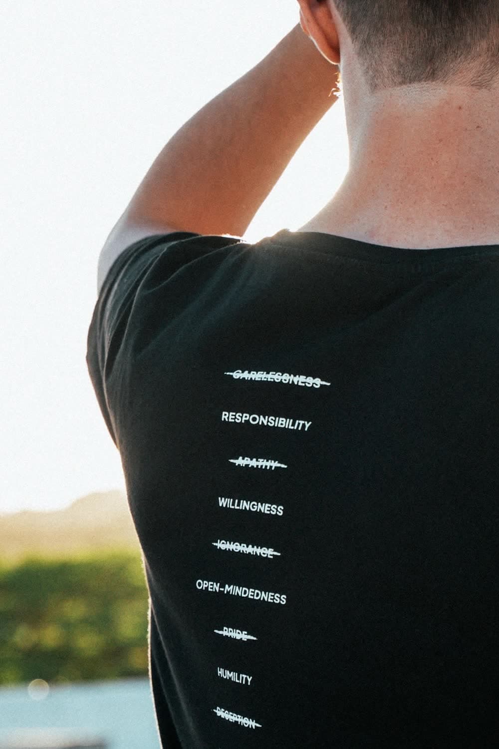 The back of a black tee in low sun: a column of struck-through words, each answered underneath by the one that replaces it