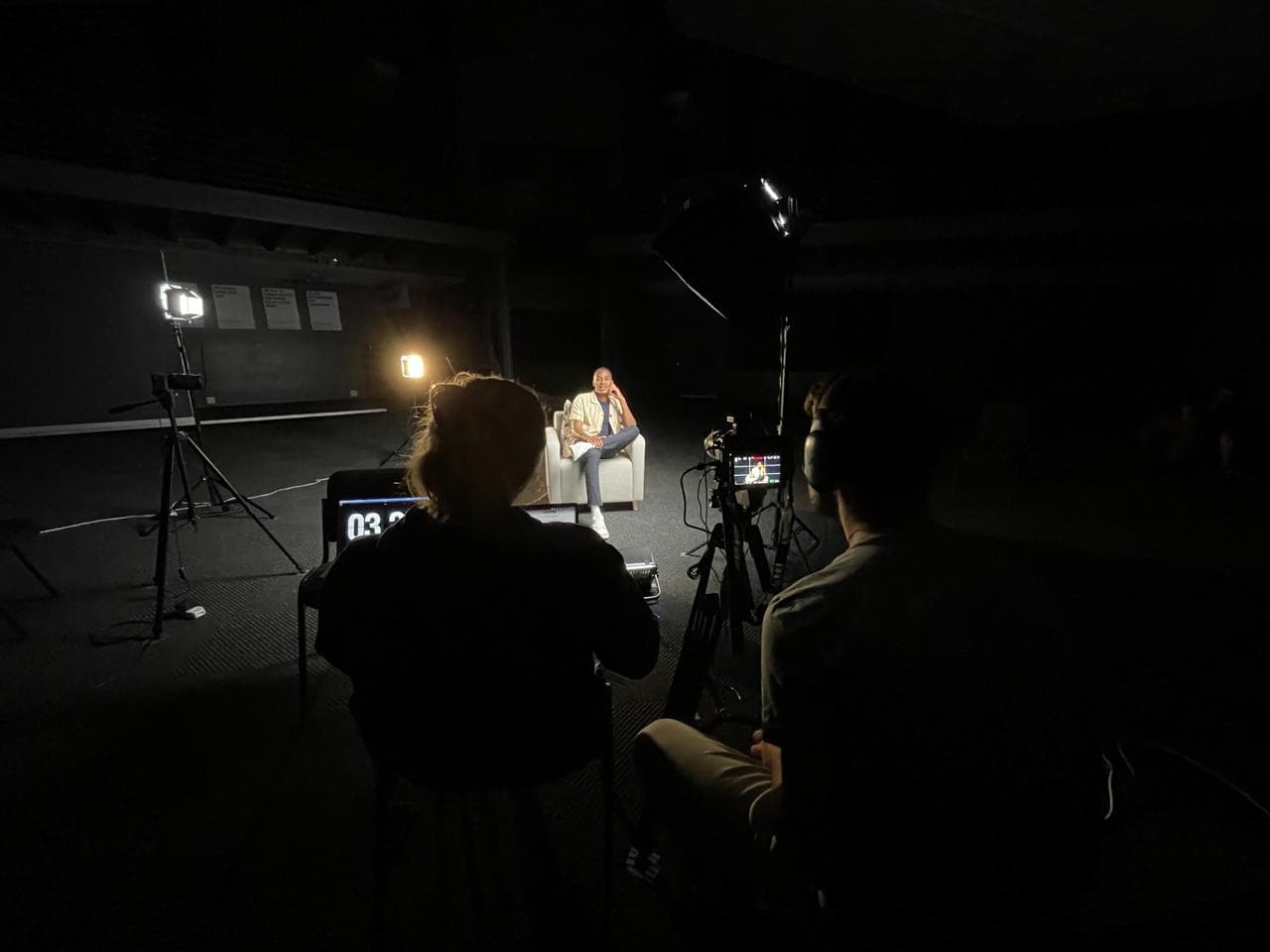 The interview set in a darkened studio: the subject lit in a chair, the crew behind camera and monitor