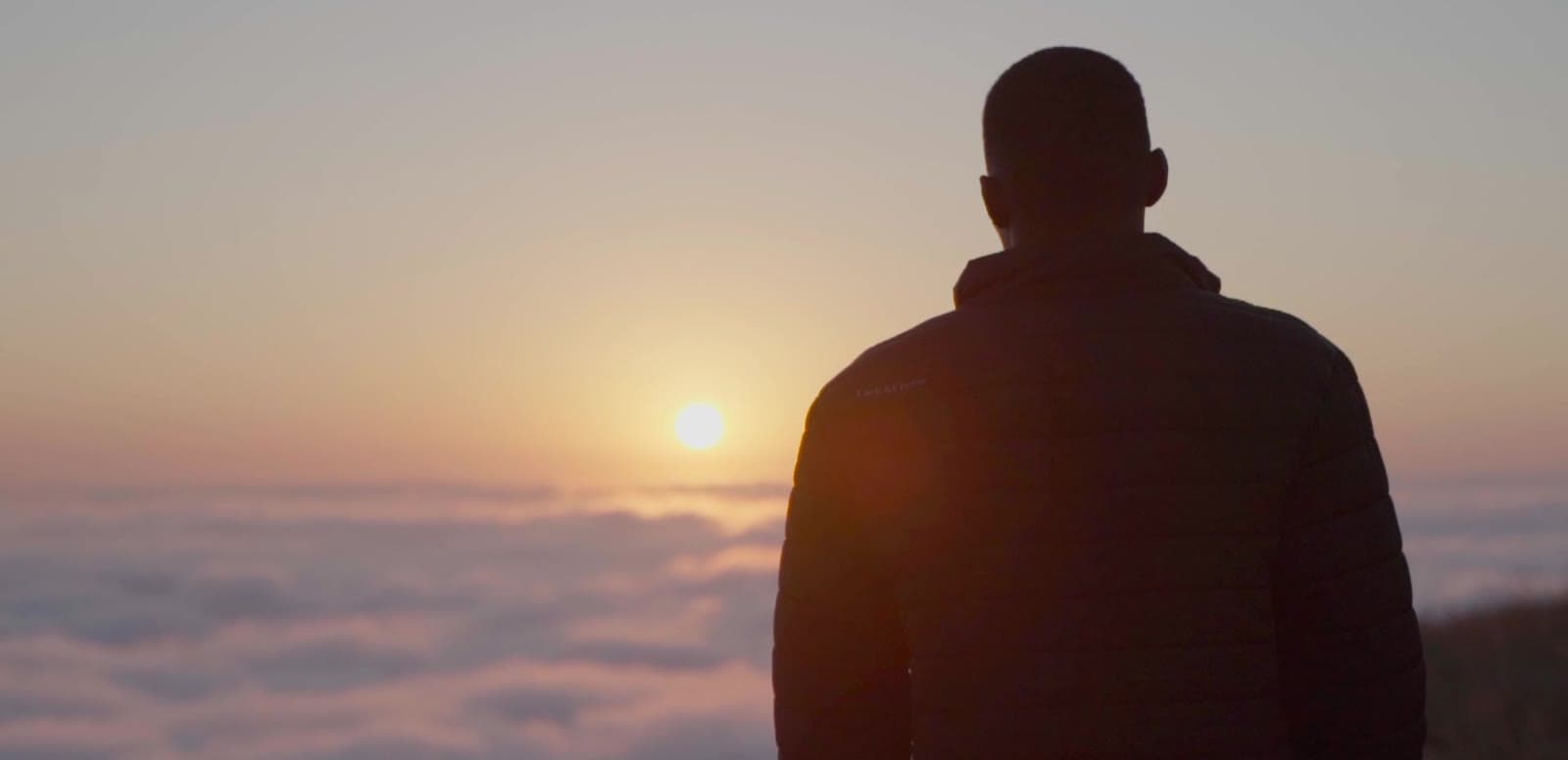 A figure standing in silhouette above a layer of cloud at sunrise